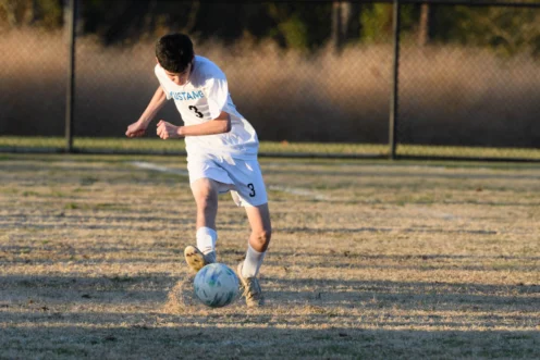 A soccer player in a white uniform with the number 3 kicks a soccer ball on a grassy field.