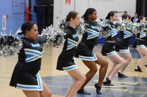A group of cheerleaders in matching black and blue uniforms perform a synchronized routine indoors, holding silver pom-poms.