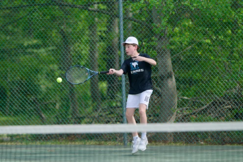 A young person in tennis attire prepares to hit a tennis ball with a racket on an outdoor court, with a green chain-link fence and trees in the background.