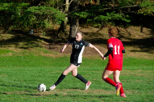 Two soccer players compete for the ball on a grassy field; one in a black uniform prepares to kick, while the other in red approaches. Trees are in the background.