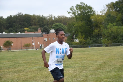 A young person wearing a race bib and a white Nike Mustang shirt runs on grass with trees and a brick building in the background.