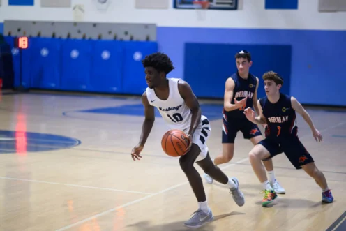 A basketball player in a white uniform dribbles the ball down the court while two players in dark uniforms defend.