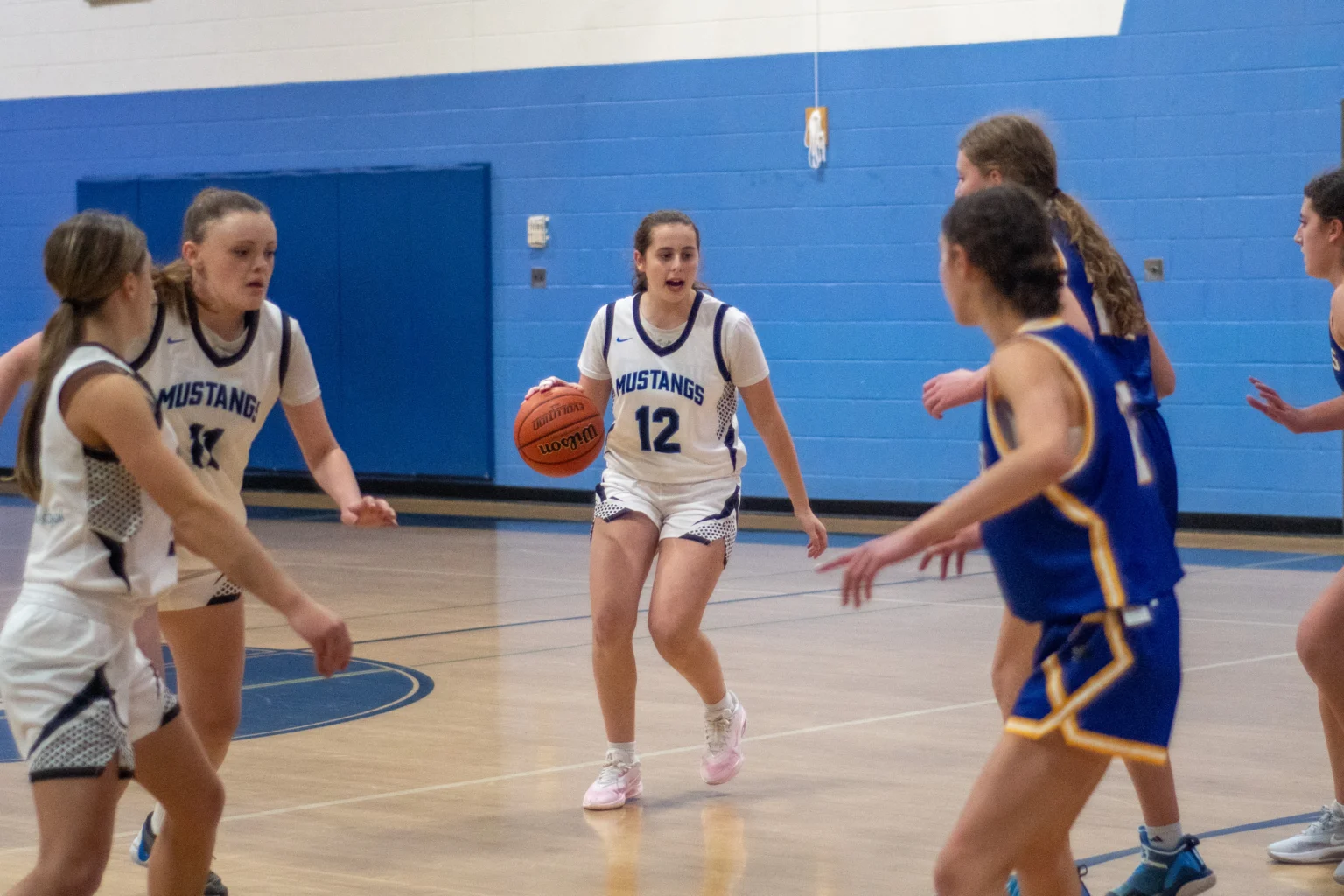 A girls’ basketball player in a white Mustangs jersey dribbles the ball while being guarded by opponents in blue uniforms during a game.