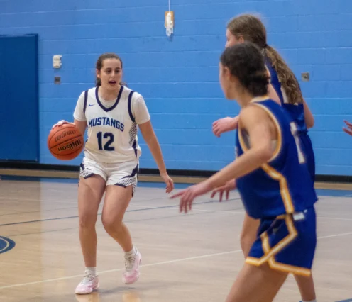 A basketball player in a white Mustangs uniform dribbles the ball while being guarded by players in blue uniforms on an indoor court.