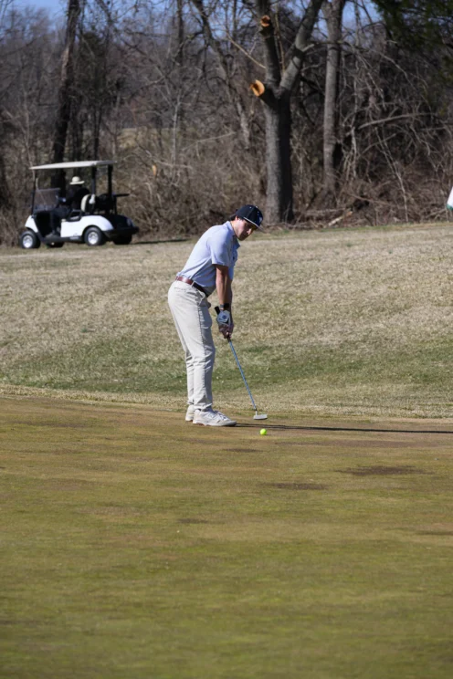 A golfer prepares to putt on a green with a golf cart and leafless trees in the background.