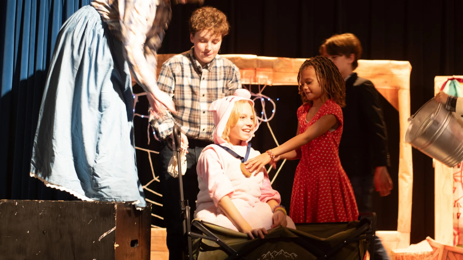 A group of children perform a play on stage, with one child in a pink pig costume sitting in a cart while others interact with her.