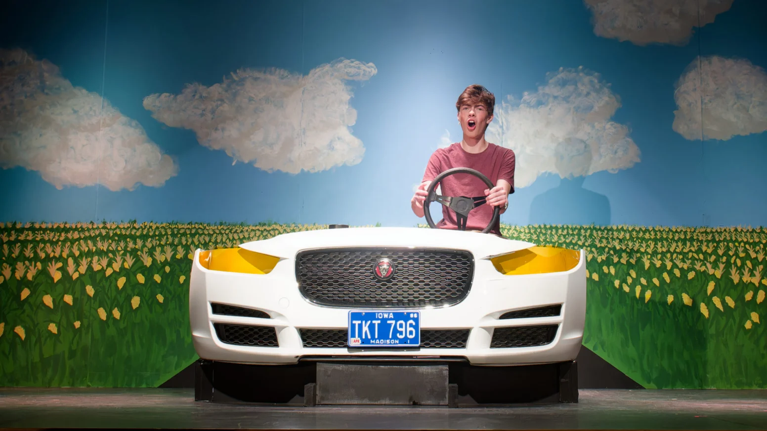 A young person sits behind the wheel of a white car prop on stage, with a painted backdrop of clouds and a field of yellow tulips.