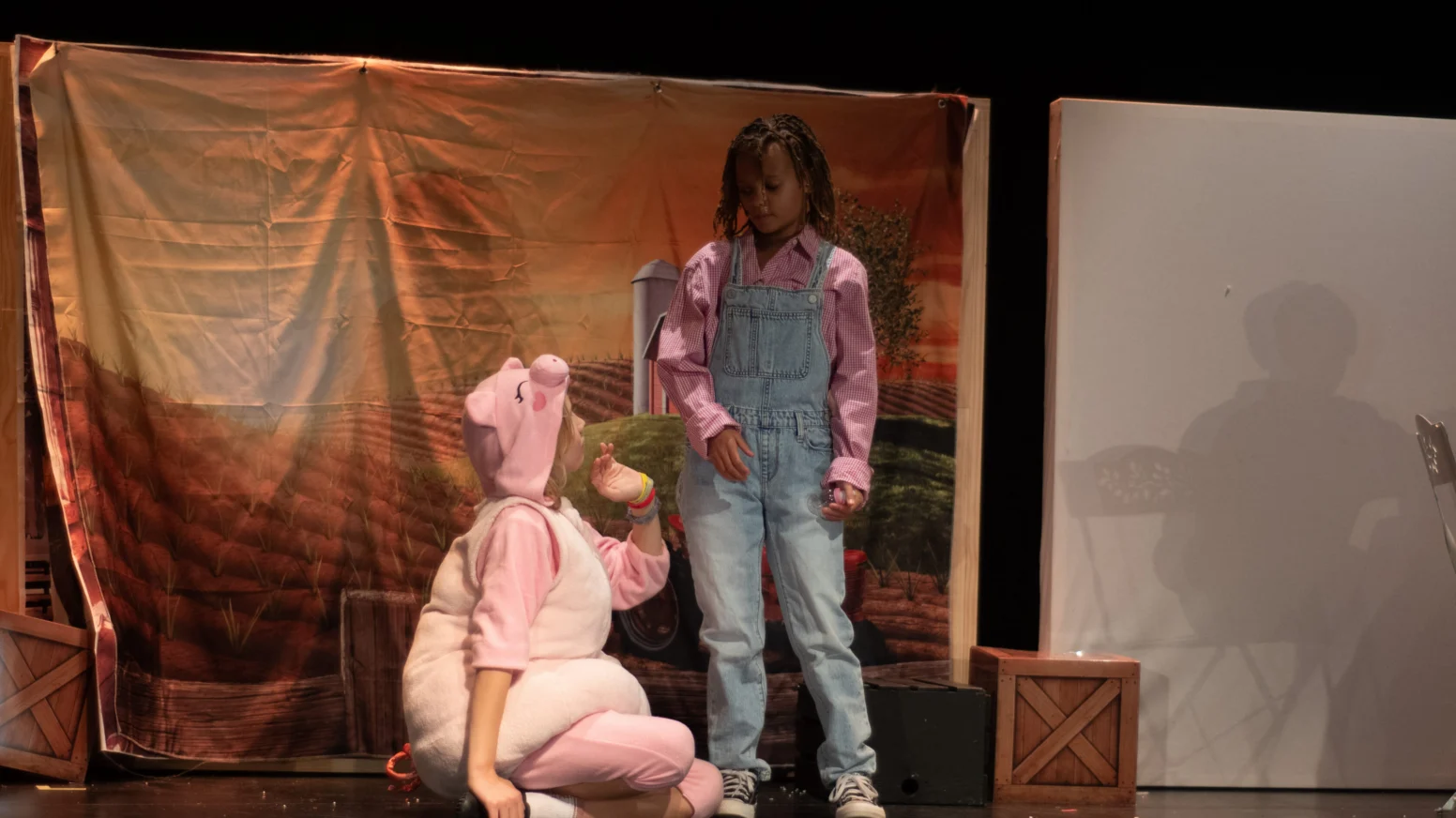 Two children perform on stage; one wears a pig costume and kneels, while the other stands in overalls and a pink shirt. A farm-themed backdrop and crates are in the background.