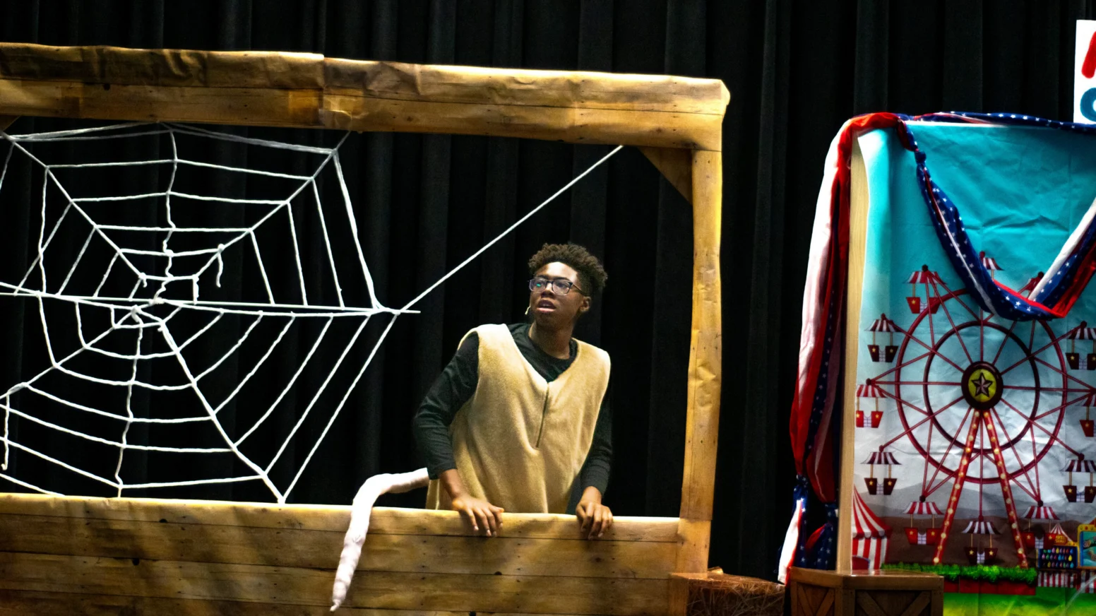 A person in costume stands behind a wooden frame with a large rope spiderweb, next to a colorful backdrop featuring a Ferris wheel.