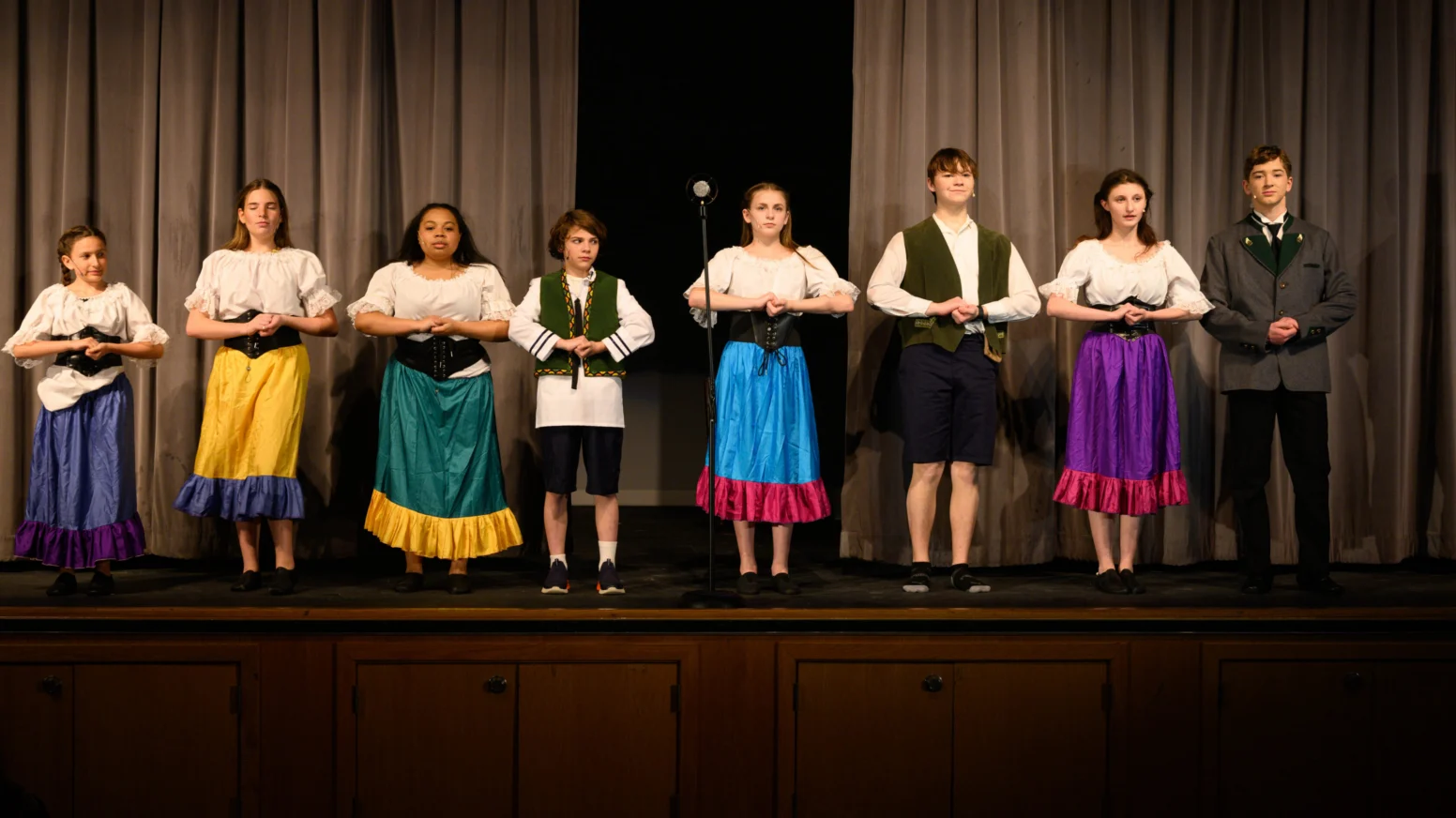 Eight young people stand onstage in costume, facing forward with hands clasped, in front of closed curtains and a microphone stand.