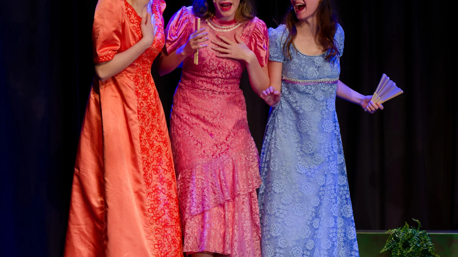 Three women in long, colorful dresses stand on stage, appearing engaged in animated conversation with expressive gestures, against a dark curtain backdrop.