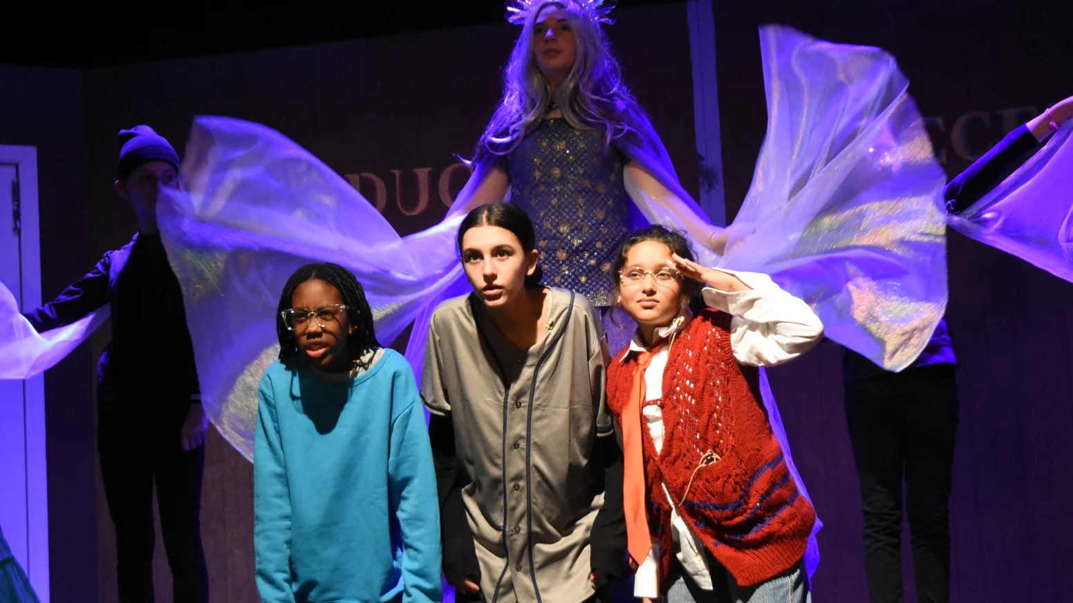 Three children kneel and pose on stage, while a figure in a long, flowing costume with a crown stands behind them with arms outstretched, under theatrical lighting.