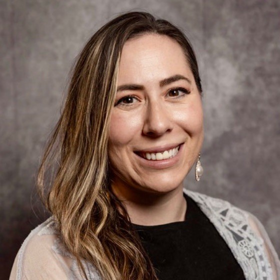 A woman with long, light brown hair and a side part smiles at the camera. She is wearing a dark top, patterned shawl, and a dangling earring. The background is gray and textured.