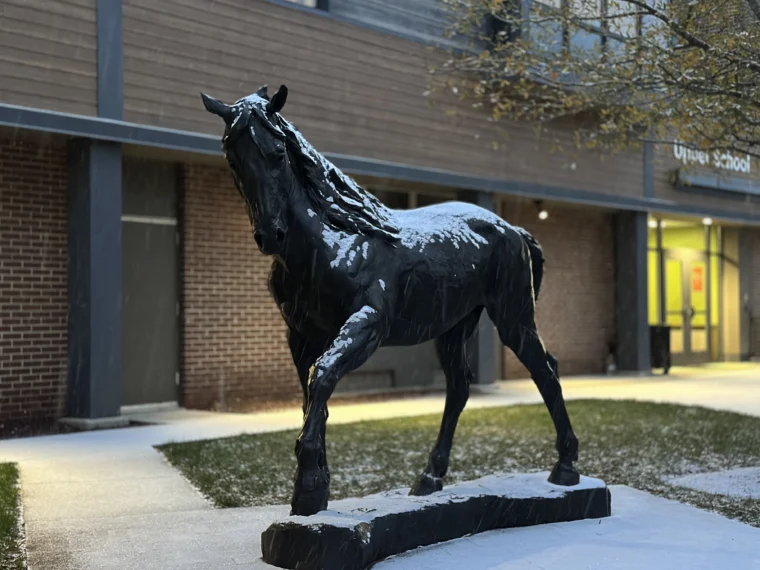 A black horse statue with a dusting of snow stands on a sidewalk in front of a brick and wood school building.