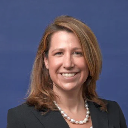 A woman with shoulder-length light brown hair, wearing a black blazer, white top, and pearl necklace, smiles in front of a solid blue background.
