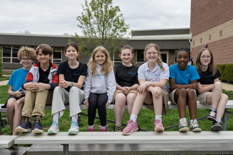Eight children sit on bleachers outside a school building, facing the camera and smiling. The group is casually dressed and the background shows grass, trees, and school walls.