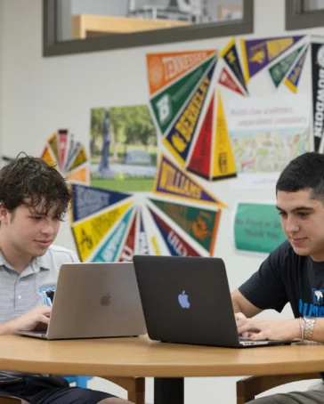 Two students sit at a round table, working on laptops in a classroom decorated with colorful college pennants on the wall.