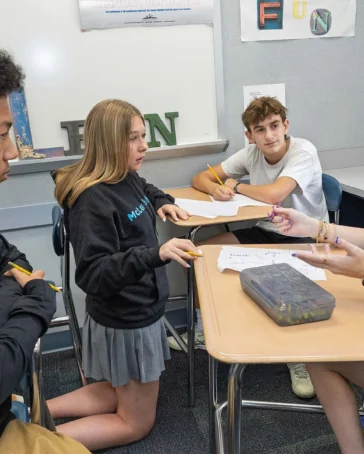 Four students sit at desks in a classroom, talking and writing on paper together during a group activity.