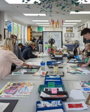 Students work on various art projects at large tables in a bright, well-equipped classroom filled with art supplies and decorations.