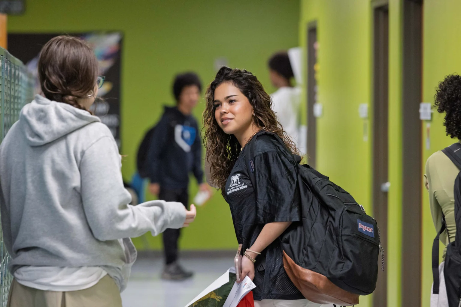 Two students talk in a hallway with green walls. One holds papers and wears a backpack; others are visible in the background.