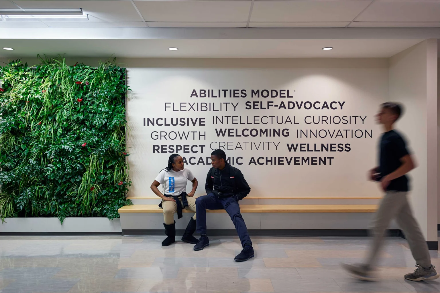 Two people sit and talk on a bench beneath a wall with motivational words, while another person walks past; a vertical garden is on the left side of the scene.