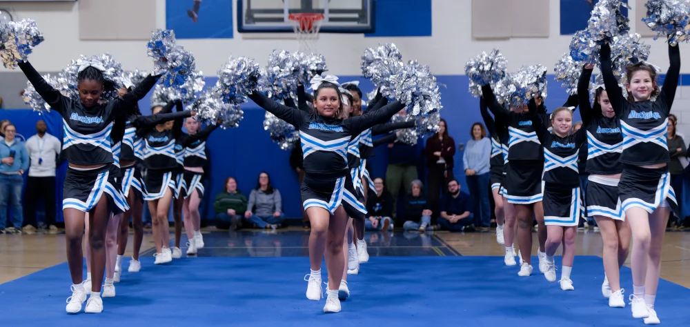 A cheerleading team in matching black and blue uniforms performs a routine indoors on a blue mat, with spectators watching in the background.