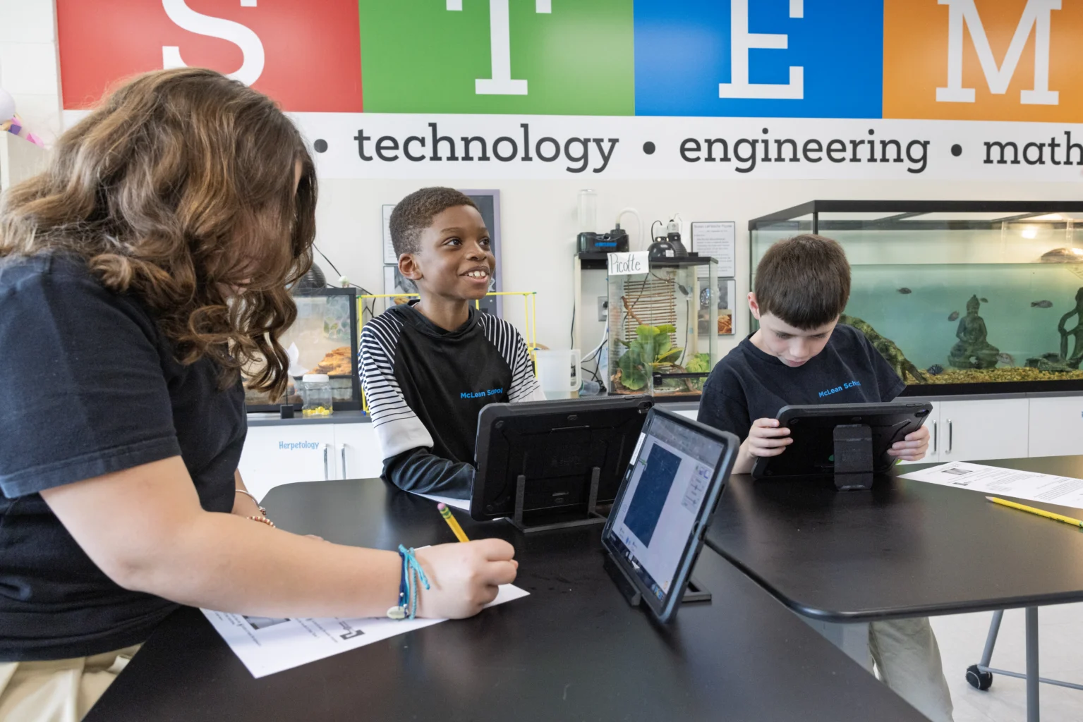 Three students sit at a table using tablets and writing, with a STEM sign and aquariums in the background.