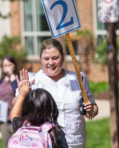 A woman holding a sign with the number 2 on a ruler high-fives a young girl with a pink backpack outdoors near a school.