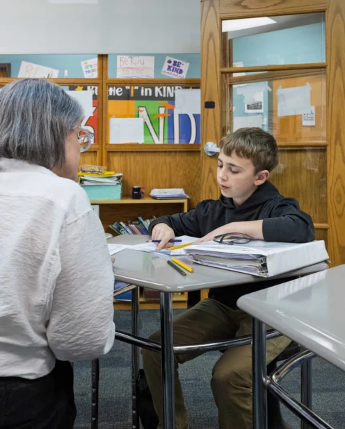 A teacher and a student sit at desks in a classroom, working together on assignments with notebooks and pencils on the table.
