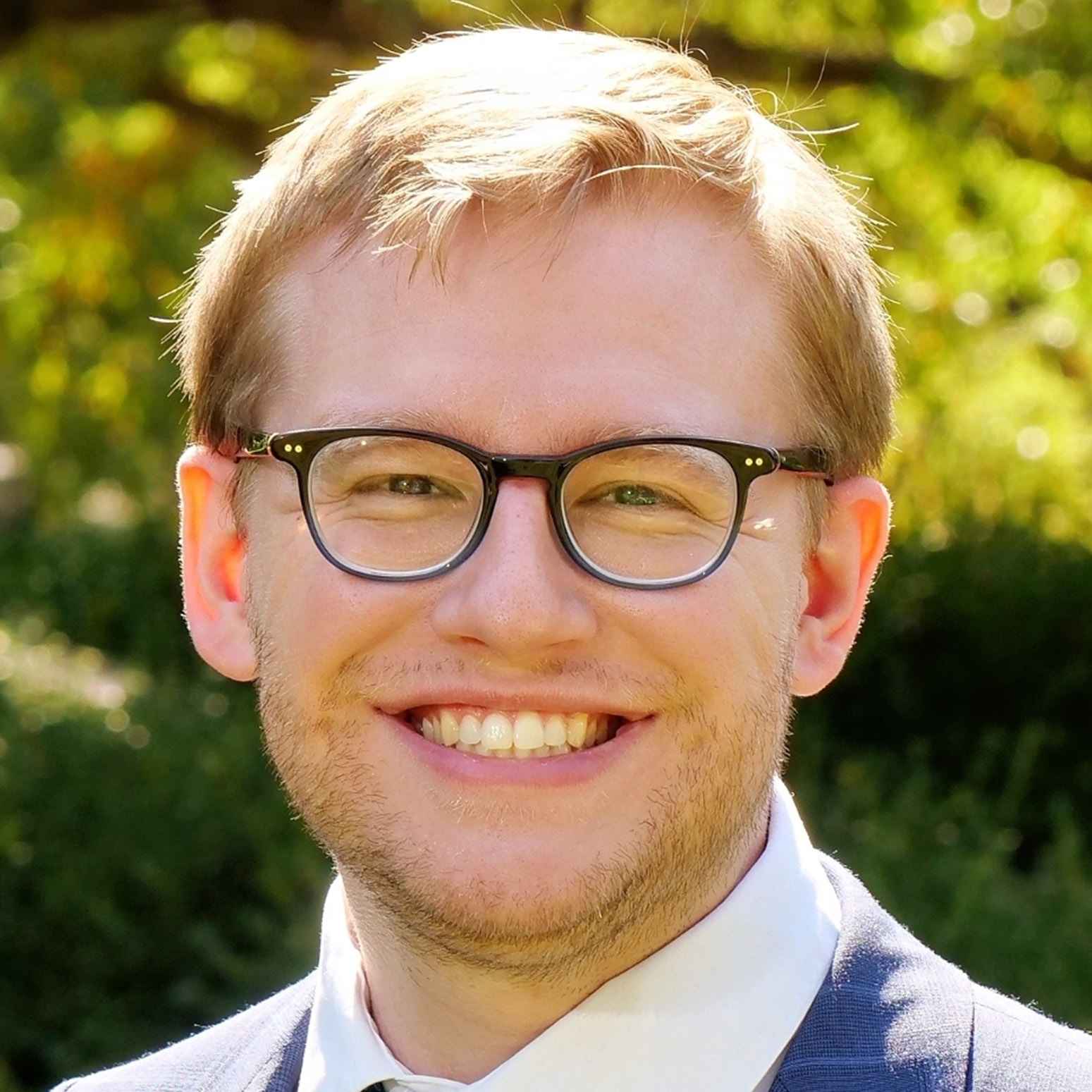 A man with short blond hair, glasses, and a light beard smiles at the camera, wearing a suit and tie outdoors with greenery in the background.