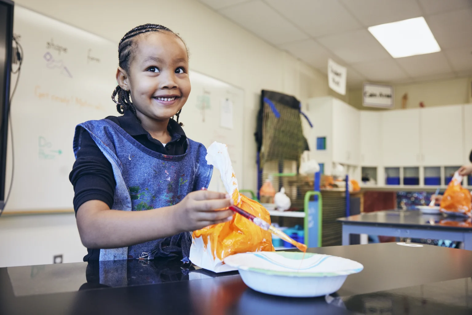 A child in an art classroom smiles while painting an object with a brush, wearing a protective apron. Art supplies and projects are visible on the table and in the background.
