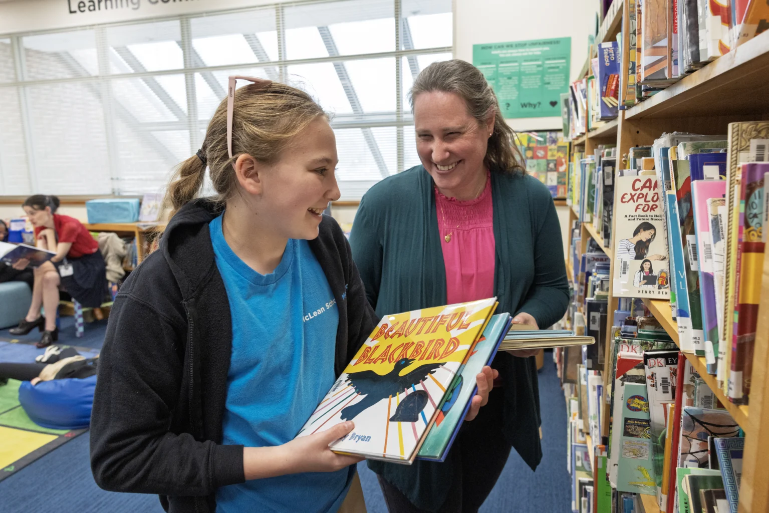 A girl and a woman smile while looking at books in a library. The girl holds a book titled "Beautiful Blackbird." Other people are visible in the background.