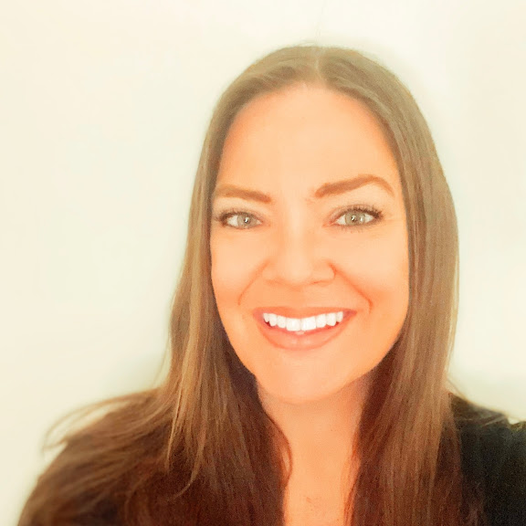 A woman with long brown hair and blue eyes smiles at the camera against a plain light background.