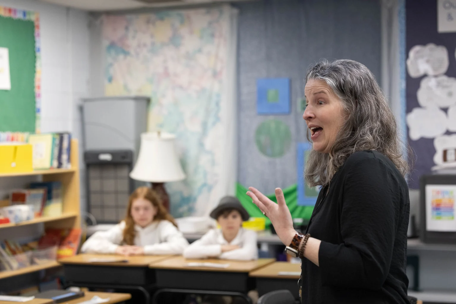 A teacher speaks to a classroom with two students seated at desks, bookshelves and educational posters visible in the background.