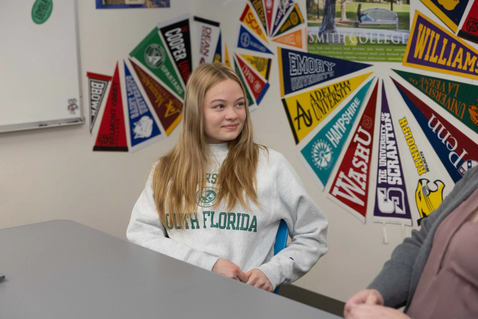 A young woman in a University of South Florida sweatshirt sits at a table in a room decorated with college pennants.