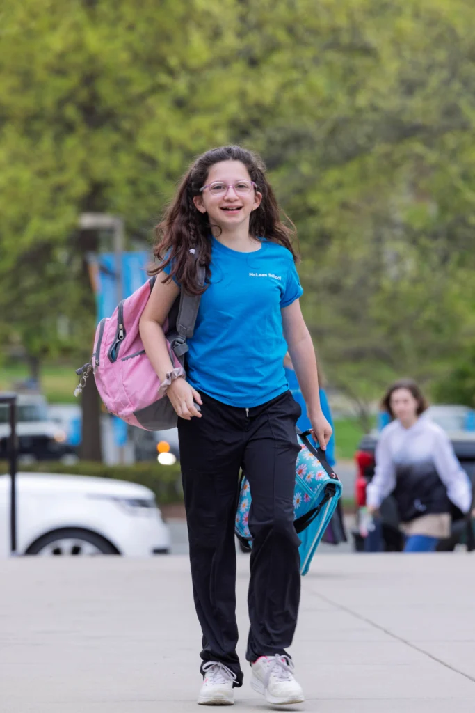 Young girl with long brown hair, wearing glasses, a blue shirt, and black pants, walks outdoors carrying a pink backpack and a blue bag. Other people and trees are in the background.