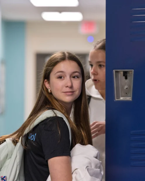Three teenage girls stand by blue lockers in a school hallway. One girl looks at the camera while the others talk to each other.