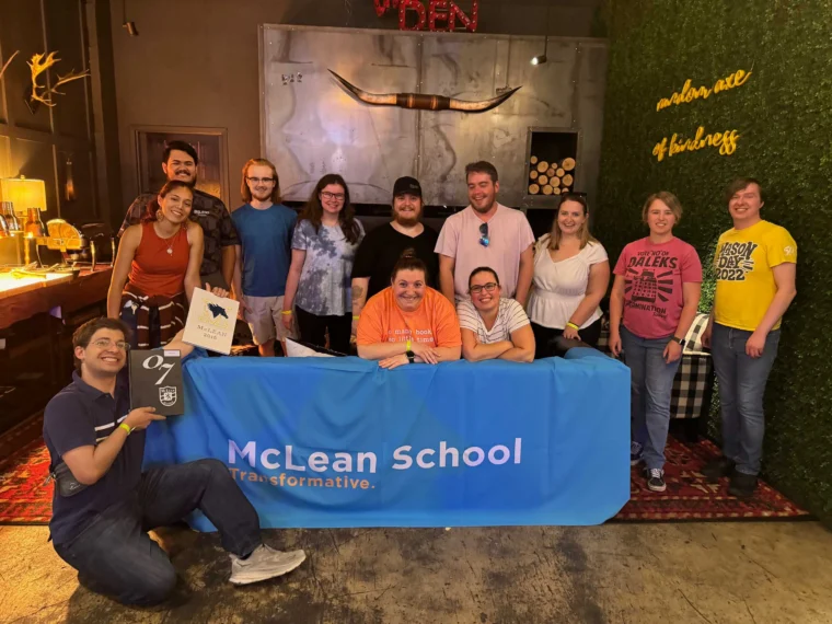 A group of people poses behind a blue McLean School table at an indoor event, with various decorations on the wall behind them.