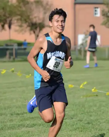 A young male runner wearing a "Mustangs" jersey and race bib 88 runs outdoors on grass during a race, with another runner and a brick building in the background.