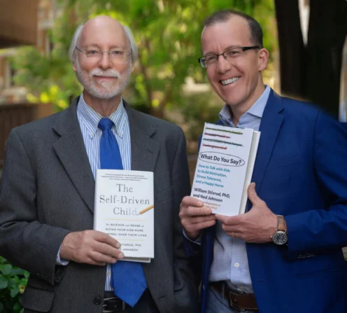 Two men in suits stand outdoors, each holding a book. The man on the left holds "The Self-Driven Child"; the man on the right holds "What Do You Say?" Both are smiling at the camera.