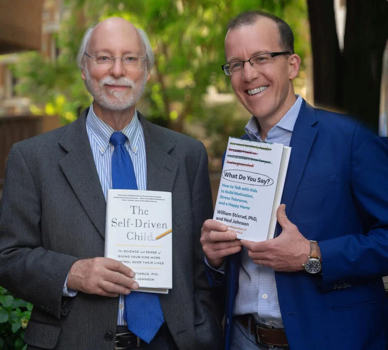 Two men in suits stand outdoors, each holding a book. The man on the left holds "The Self-Driven Child"; the man on the right holds "What Do You Say?" Both are smiling at the camera.