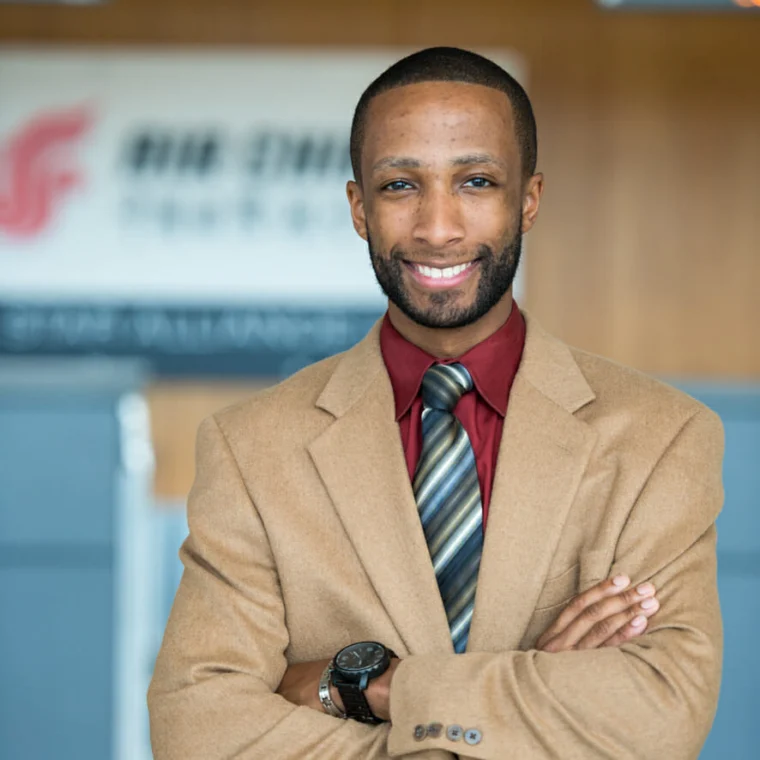 Man wearing a tan blazer, red shirt, and striped tie stands with arms crossed and smiles at the camera; blurred office background with a logo sign.