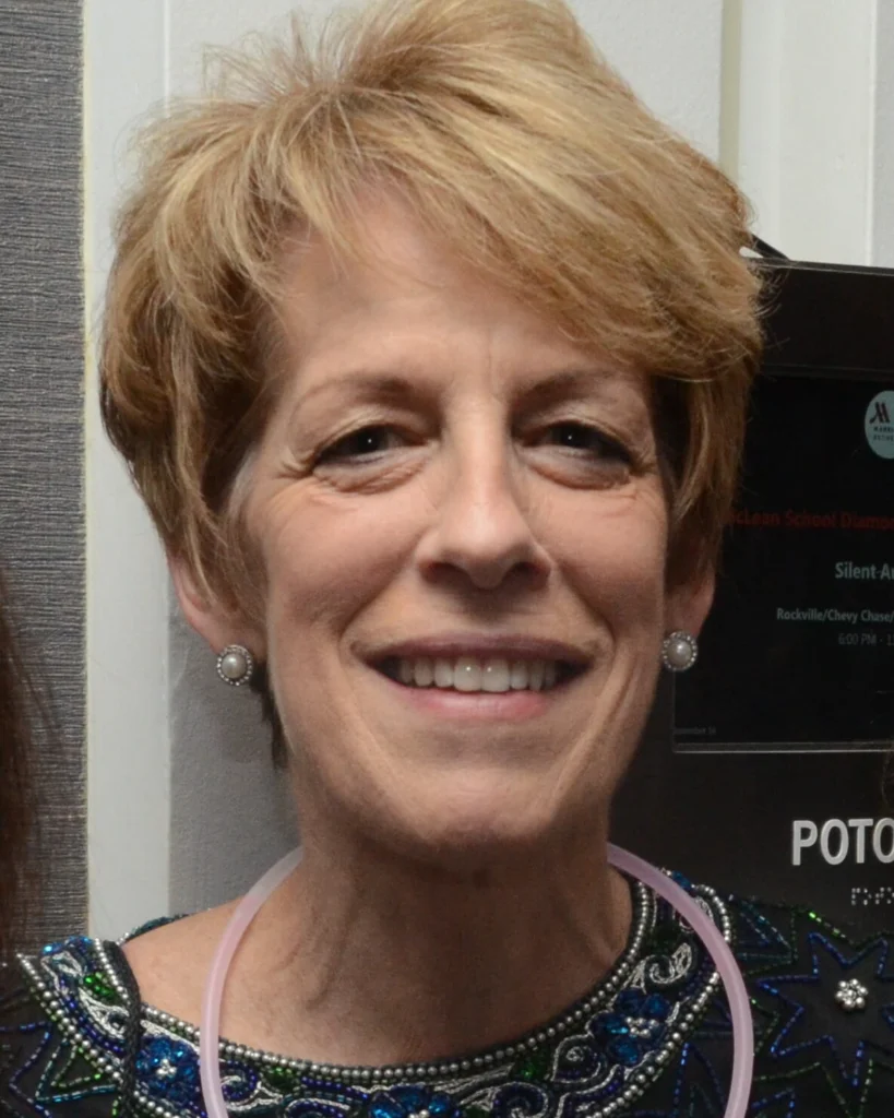 A woman with short light brown hair, wearing pearl earrings and a sequin-patterned top, is smiling at the camera.