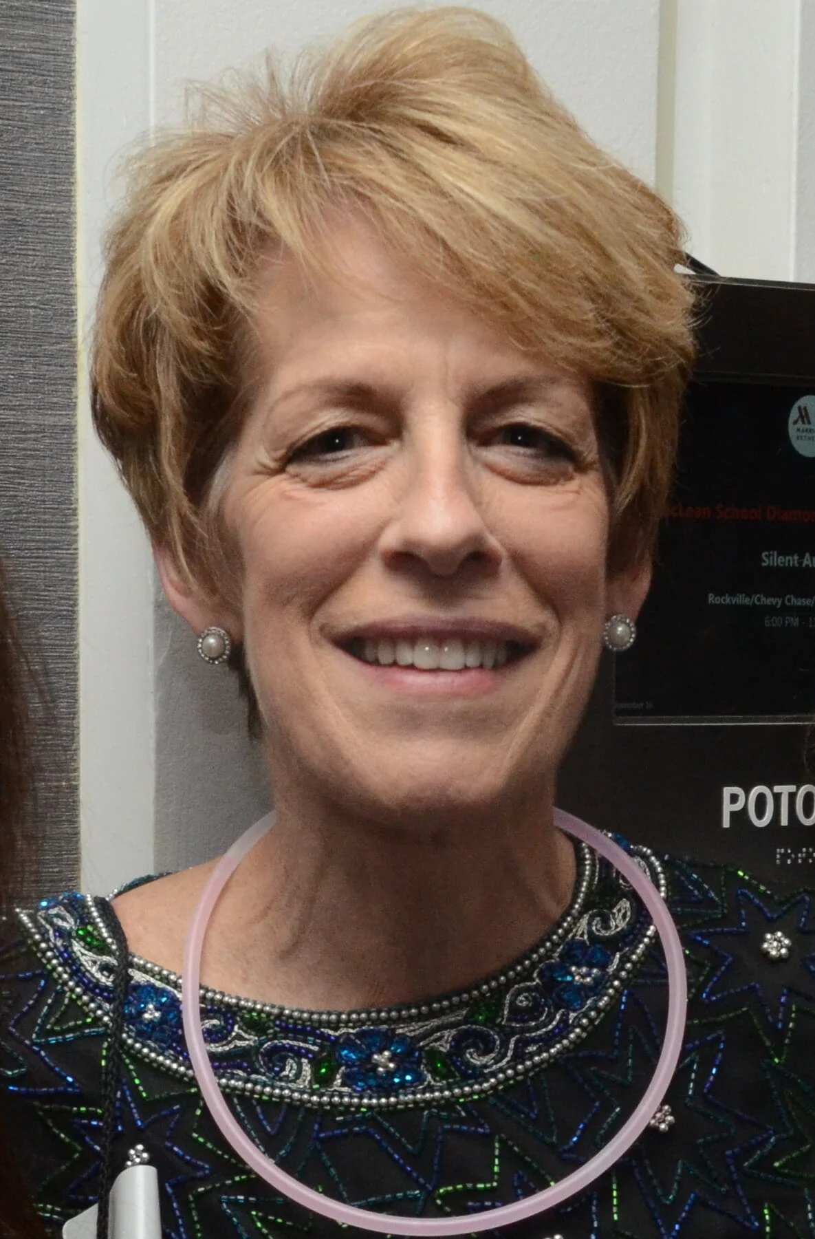 A woman with short blonde hair, wearing pearl earrings, a dark patterned top, and a light pink glow necklace, smiles at the camera.