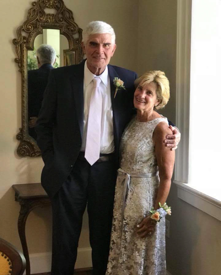 An older man in a suit and an older woman in a silver dress stand together indoors, smiling, wearing corsages, with a decorative mirror and window in the background.