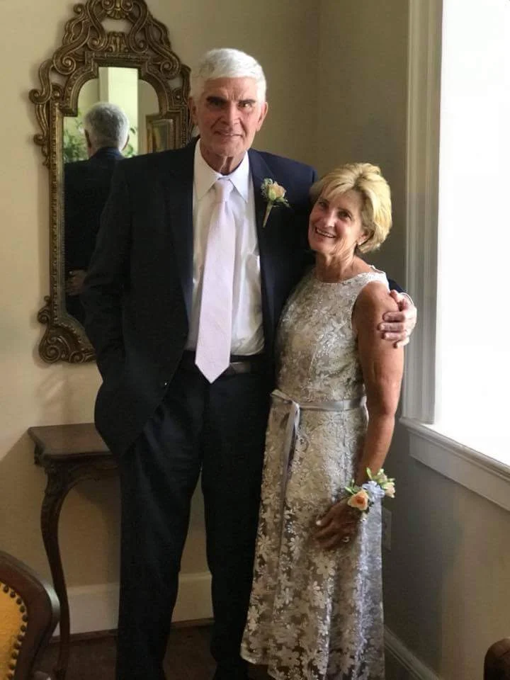 An older man in a suit and an older woman in a lace dress stand together, smiling, in a well-lit room with a mirror and table in the background.