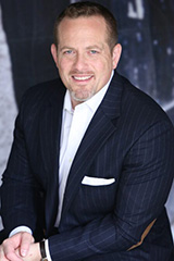 A man in a dark pinstripe suit and white shirt sits, smiling, with a blurred black and white background behind him.