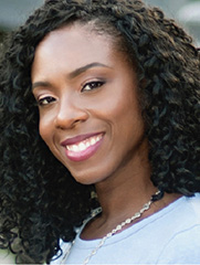 Woman with curly black hair and a light purple top smiling at the camera in an outdoor setting.