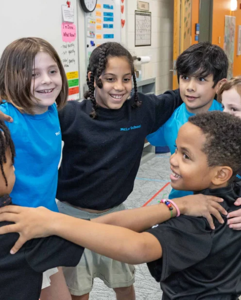 Six young children stand in a circle with their arms around each other, smiling, in a classroom setting with educational posters and shelves in the background.