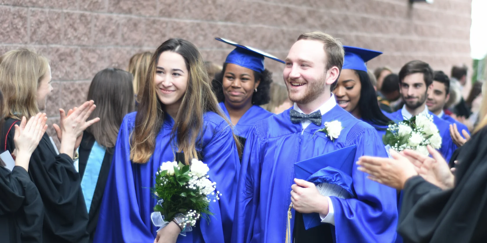 Graduates in blue caps and gowns walk in a procession, holding flowers and diplomas, while people on the sides applaud.